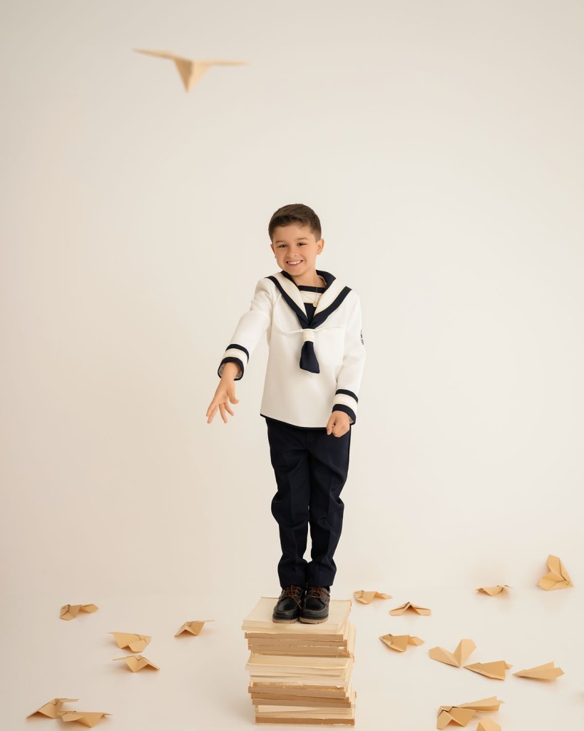 Fotografías de comunión en Gijón: niño vestido de marinero lanzando aviones de papel sobre una pila de libros